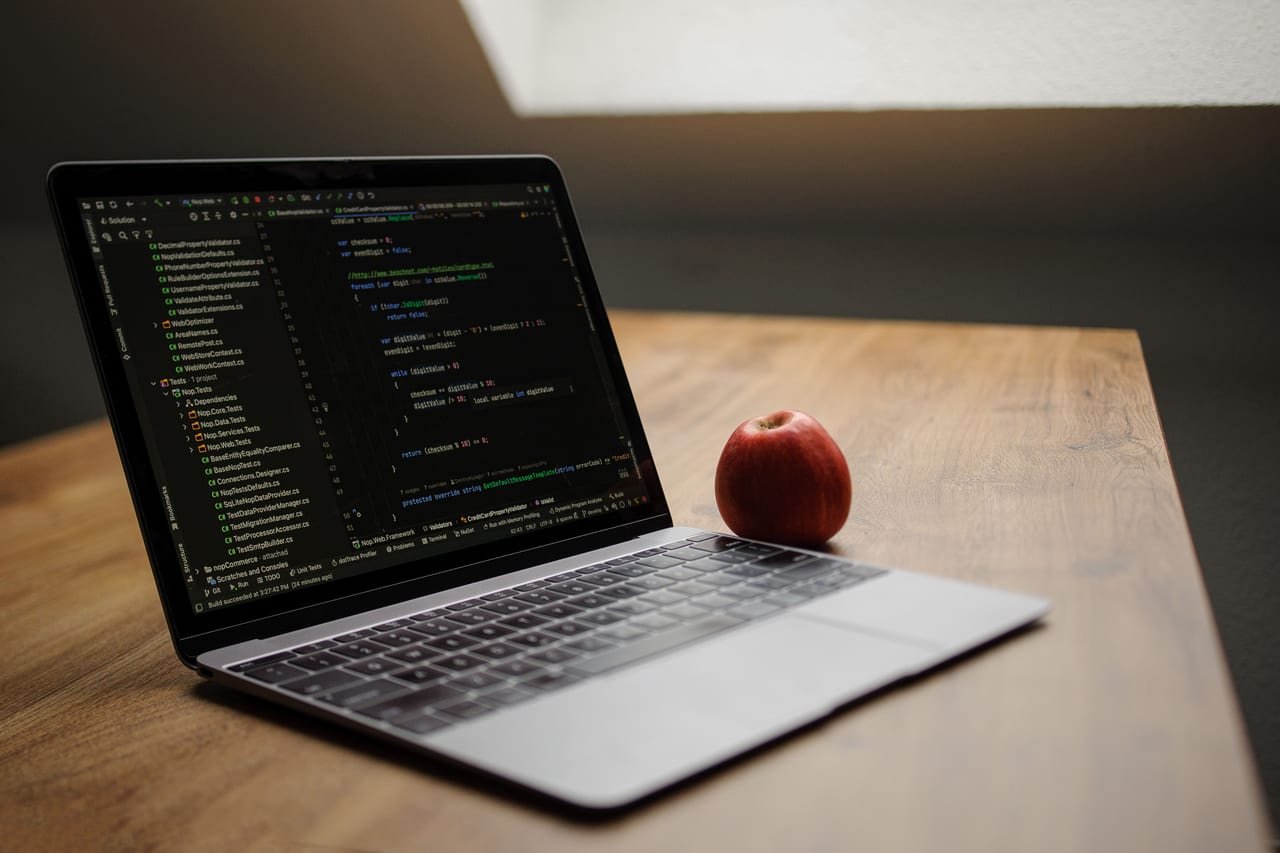 Aufgeklapptes Notebook mit Quelltext auf einem Holztisch, daneben liegt ein roter Apfel